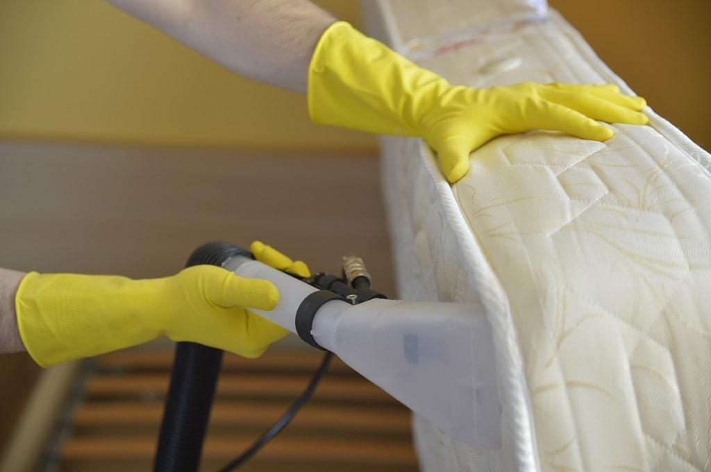 ndis-mattress-cleaning-fc ndis mattress cleaner wearing yellow rubber gloves, holding a mattress up with one hand and using a machine to clean it with the other